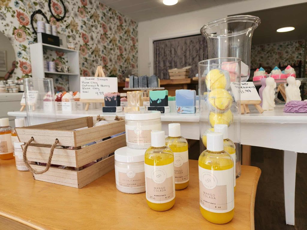 Brightly packaged bath bombs, lotions, and soaps on a wooden table, with floral wallpaper and display shelves in the background.