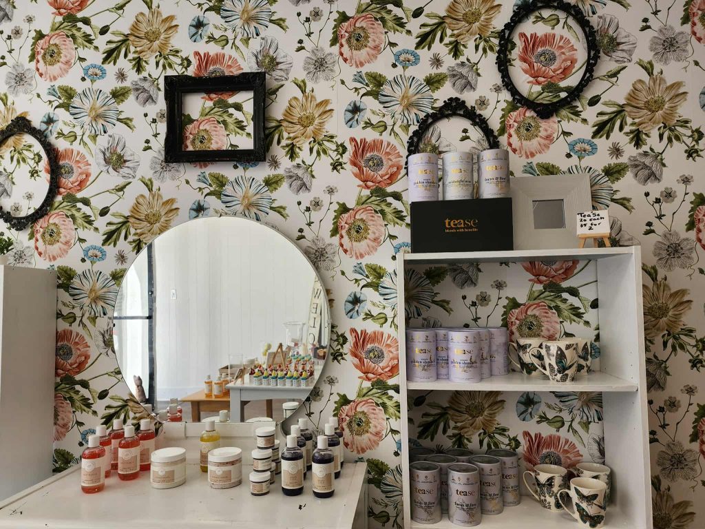Brightly packaged bath bombs, lotions, and soaps on a wooden table, with floral wallpaper and display shelves in the background.