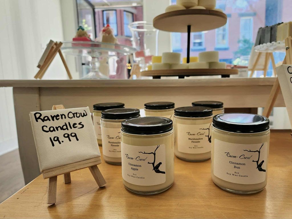 A display of Raven Crow soy candles in scents like Cinnamon Bun and Marshmallow Fireside, set against a window with historic Uptown buildings outside.