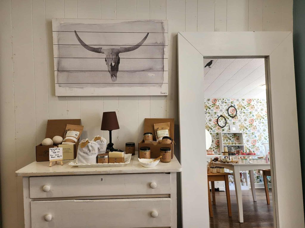 Raven Crow in Uptown Saint John, A rustic white dresser topped with bath products, candles, and gift sets, with a bull skull painting above and floral wallpaper reflected in a mirror.