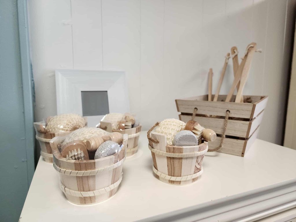 Wooden pails filled with natural bath and spa accessories, neatly arranged on a white shelf with more self-care tools in a crate nearby.