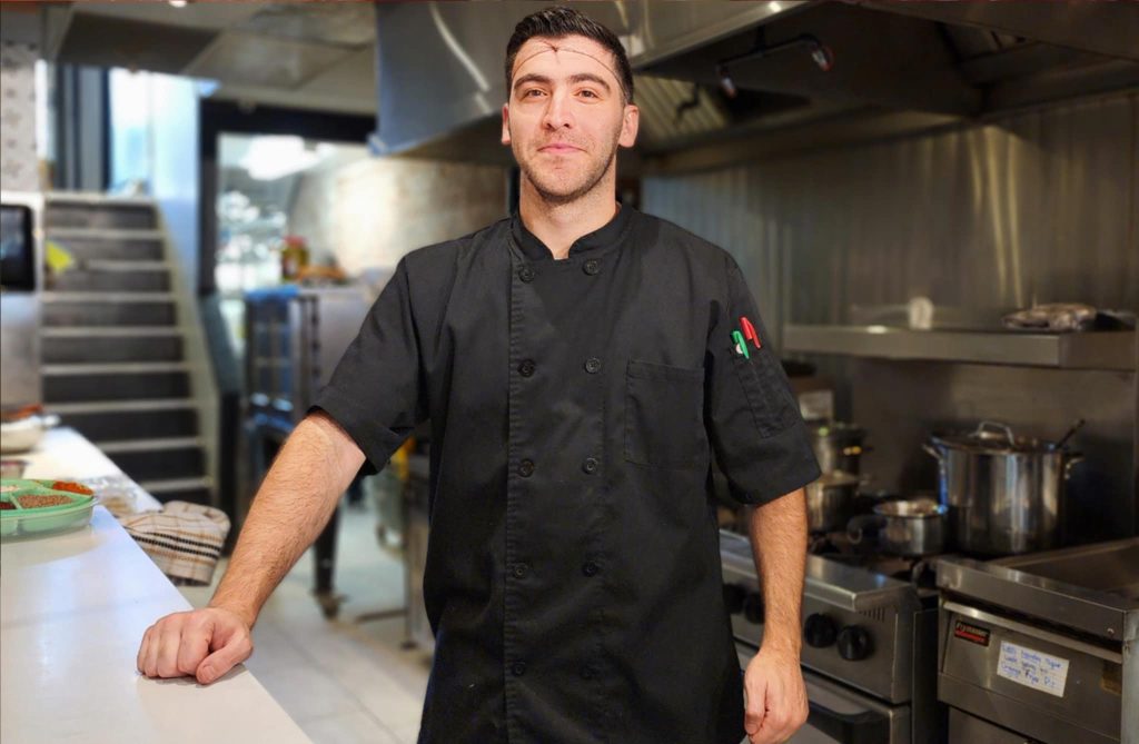 Chef Ugur Postalli standing in the kitchen of Sumac & Thyme, wearing a black chef’s coat, with cooking pots and patterned tiles in the background.