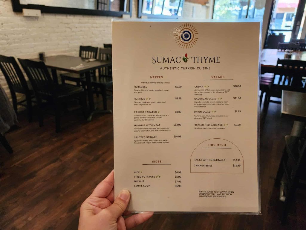 A hand holding the Sumac & Thyme menu inside the restaurant, with wooden tables, chairs, and exposed brick walls in the background.