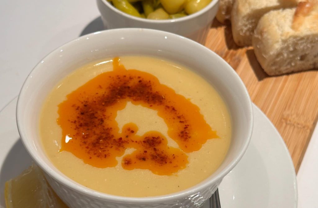 A white bowl of Turkish lentil soup topped with red chili oil, served with fresh bread and green peppers on a wooden board.