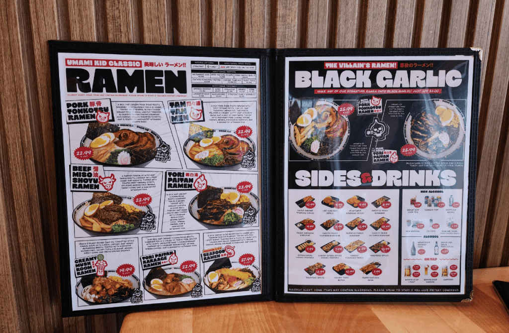 The Umami Kid menu displayed on a wooden table in Uptown Saint John, highlighting signature ramen options, sides, and Japanese drinks.