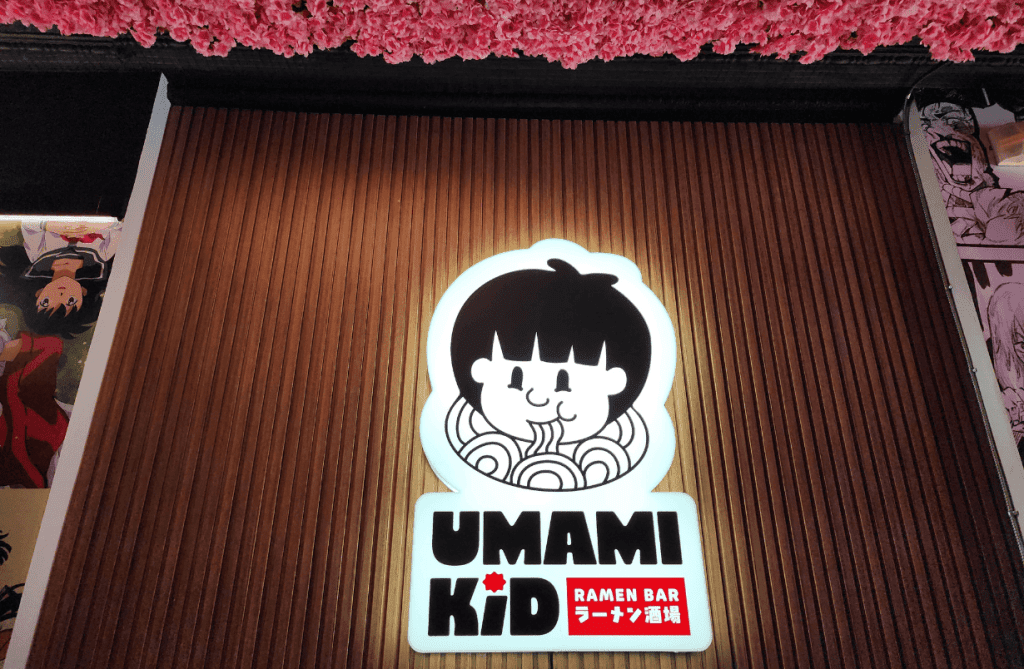 The glowing Umami Kid ramen bar logo sign inside the Uptown Saint John restaurant, framed by warm wood paneling and pink flower ceiling décor.