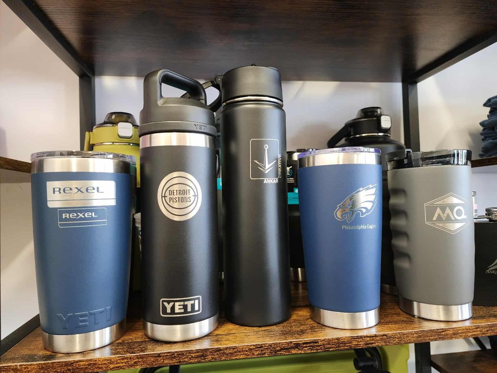 Assorted engraved tumblers and water bottles with custom logos on a wooden shelf inside Ankar Creative.