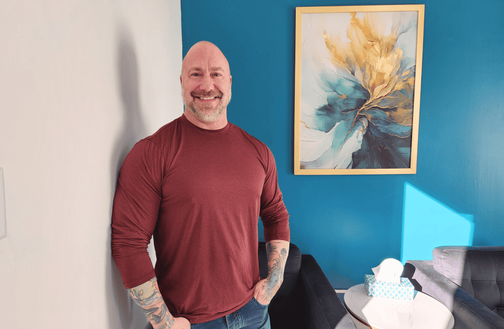 Portrait of Randy Richard, owner of Haircraft Studio, smiling inside his Uptown Saint John salon beside a teal feature wall and artwork.