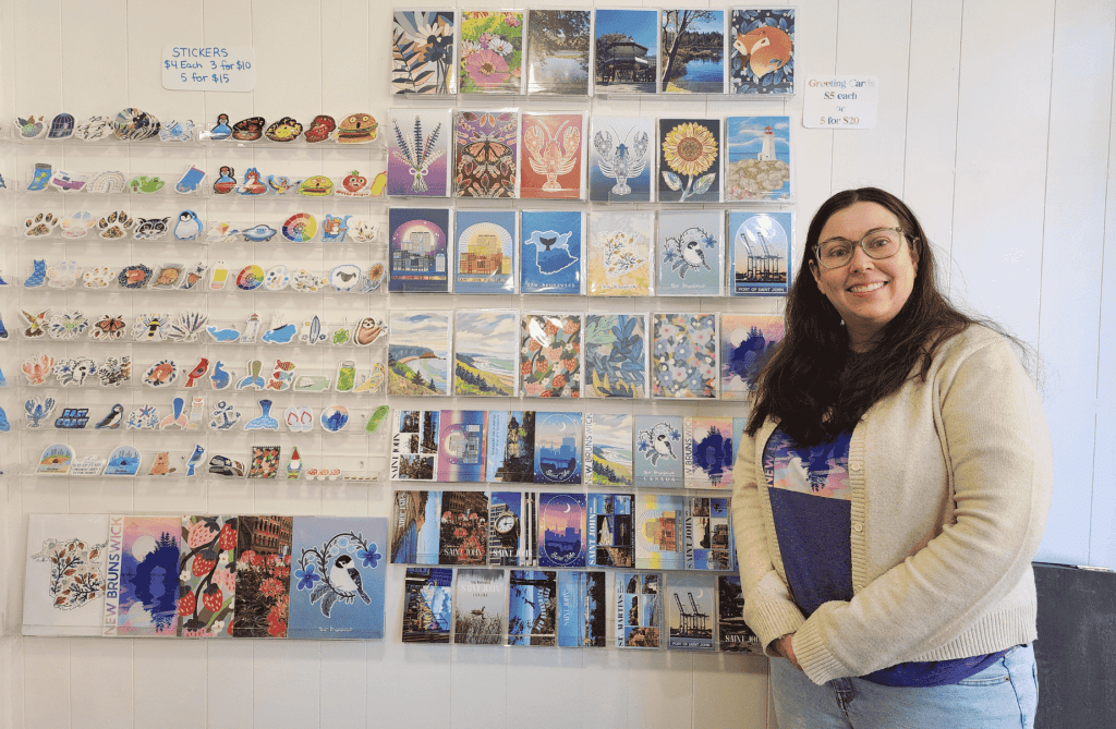 Owner Monica Memory stands inside Memory Made Gifts in Uptown Saint John beside a display wall of illustrated stickers, greeting cards, and locally inspired artwork.