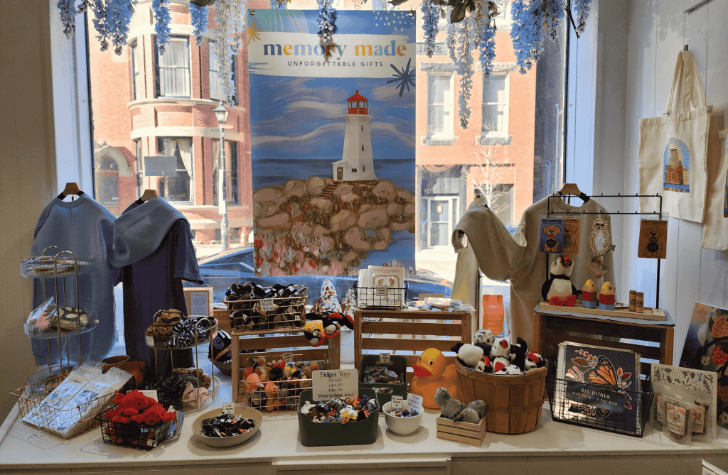 Front window display at Memory Made Gifts featuring a lighthouse illustration, apparel, and curated Canadian-made gifts in Uptown Saint John.