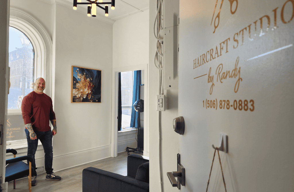 Owner Randy Richard standing inside Haircraft Studio in Uptown Saint John, a bright, welcoming salon with tall windows and historic street views.